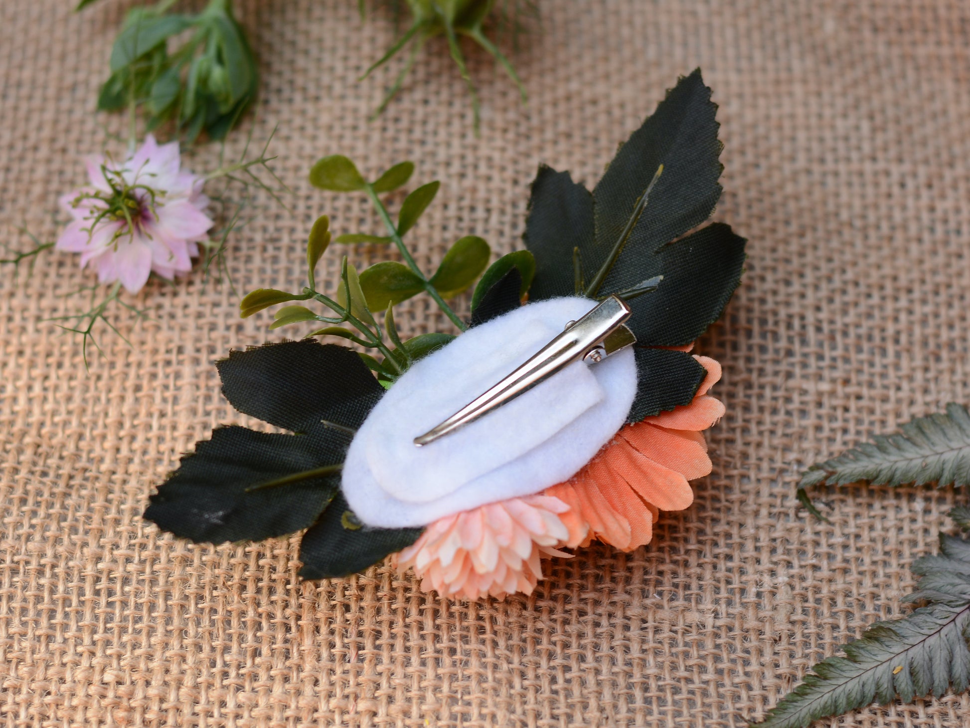 A fabric flower hair clip accessory featuring shown upside down with a white felt base and a silver metal hair clip, displayed on a burlap background.