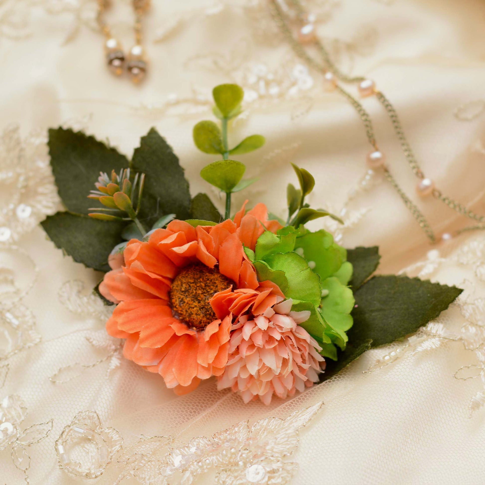 An artificial floral hair accessory featuring a prominent orange daisy, a light pink flower and green leaves displayed on a cream lace fabric background.