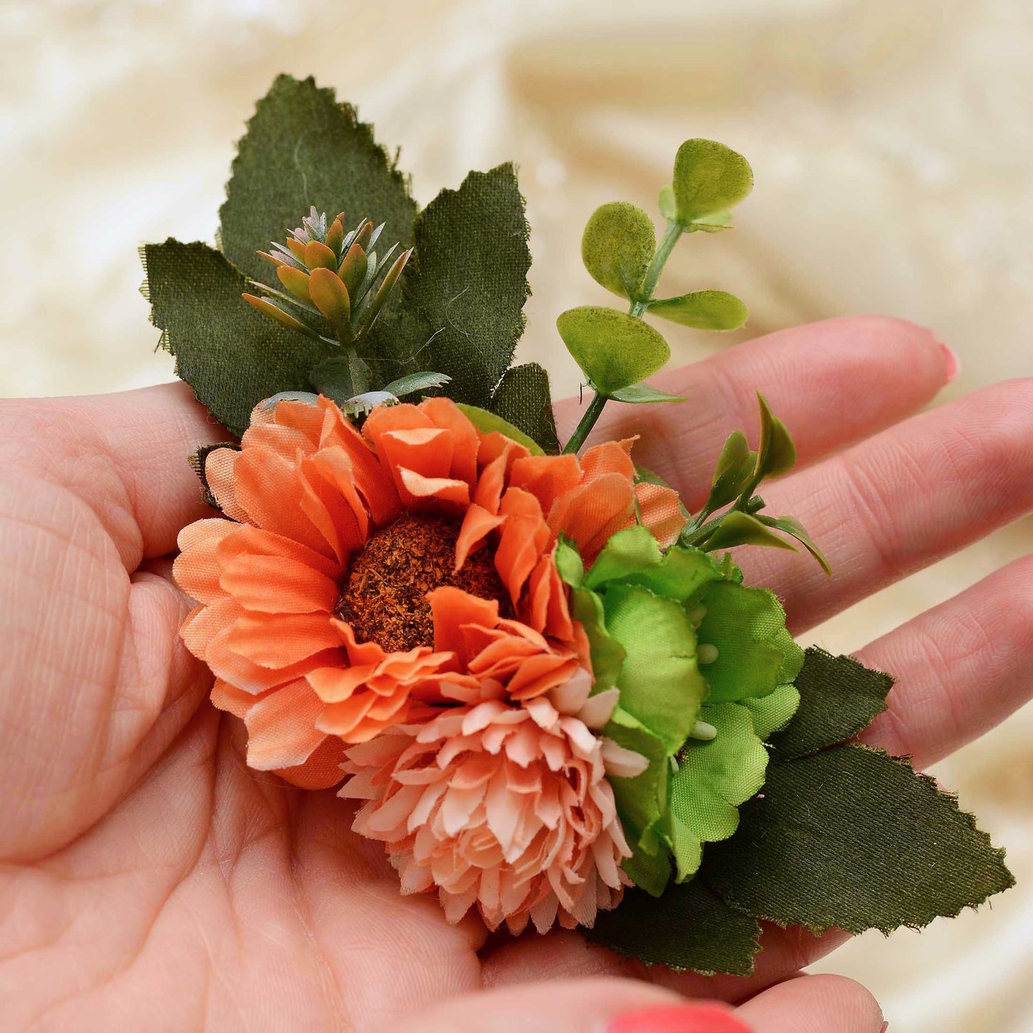 An artificial flower hair clip with orange, peach, and green flowers and leaves, held in a person's hand against a soft beige fabric background.