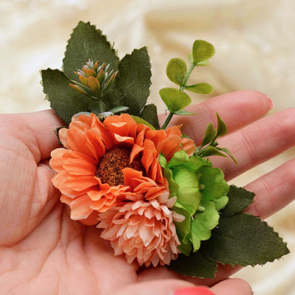 An artificial flower hair clip with orange, peach, and green flowers and leaves, held in a person's hand against a soft beige fabric background.