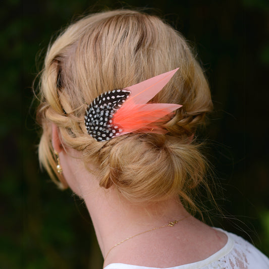 Person with a braided low bun hair style featuring a decorative coral pink feather accessory against a dark background