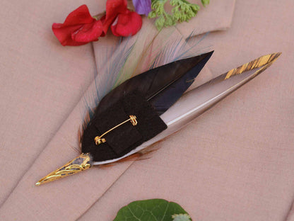 A decorative feather pin with a black feather, gold-toned metal tips, and a gold safety pin clasp, displayed on a beige fabric background with red and green floral accents.