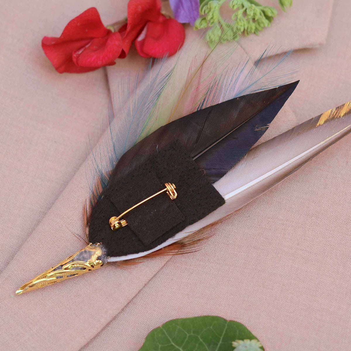 A decorative feather pin with a black feather, gold-toned metal tips, and a gold safety pin clasp, displayed on a beige fabric background with red and green floral accents.