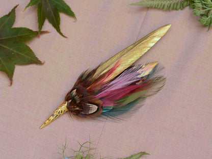 A decorative feather brooch with a gold-toned pointed metal tip and gold-painted and multicoloured feathers, displayed on a beige fabric background with small red and purple flowers nearby.