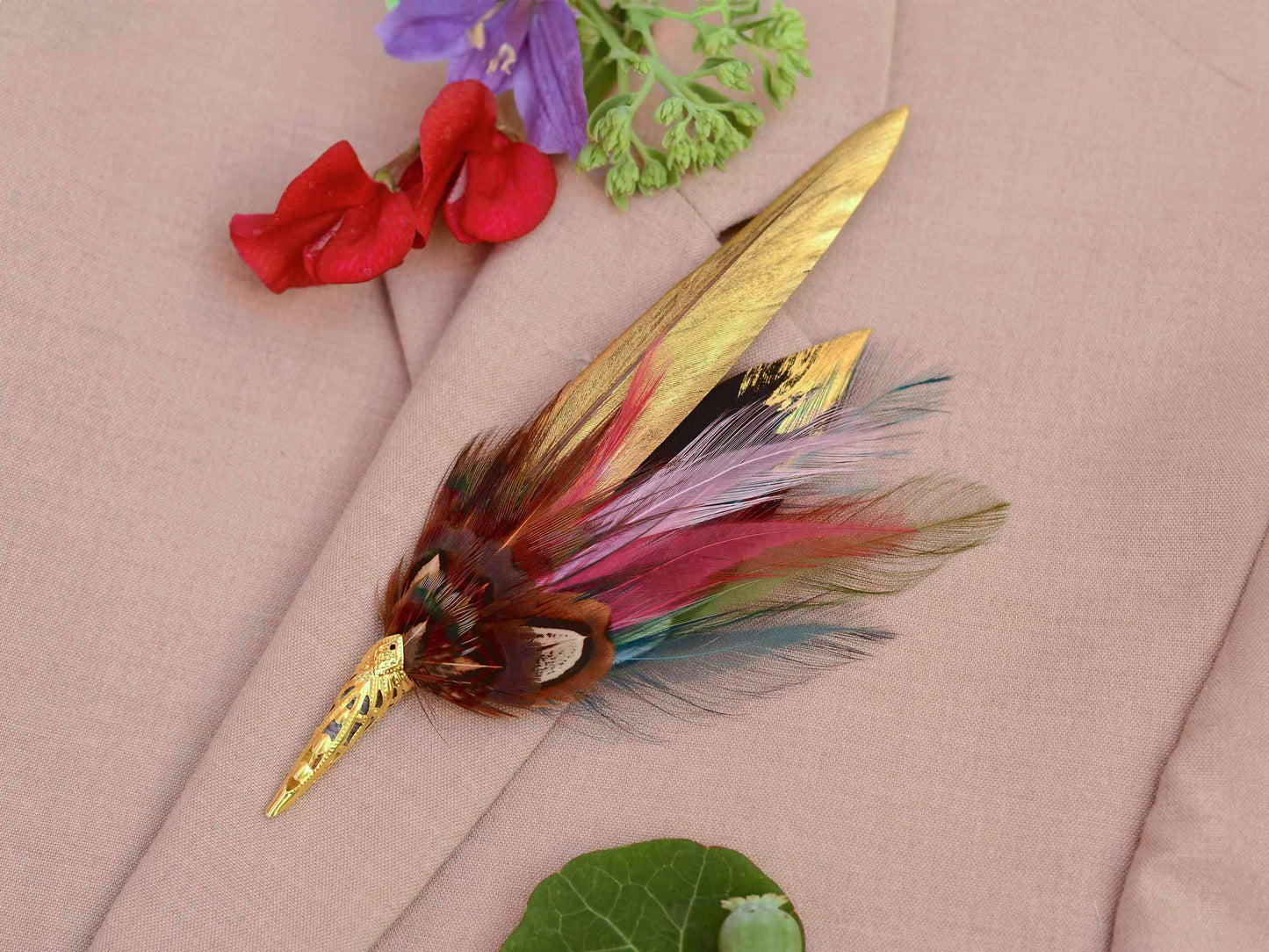 A decorative feather brooch with a gold-toned pointed metal tip and gold-painted and multicoloured feathers, displayed on a beige fabric background with small red and purple flowers nearby.