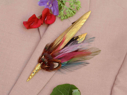 A decorative feather brooch with a gold-toned pointed metal tip and gold-painted and multicoloured feathers, displayed on a beige fabric background with small red and purple flowers nearby.