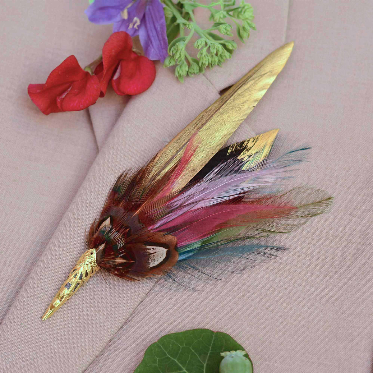 A decorative feather brooch with a gold-toned pointed metal tip and gold-painted and multicoloured feathers, displayed on a beige fabric background with small red and purple flowers nearby.