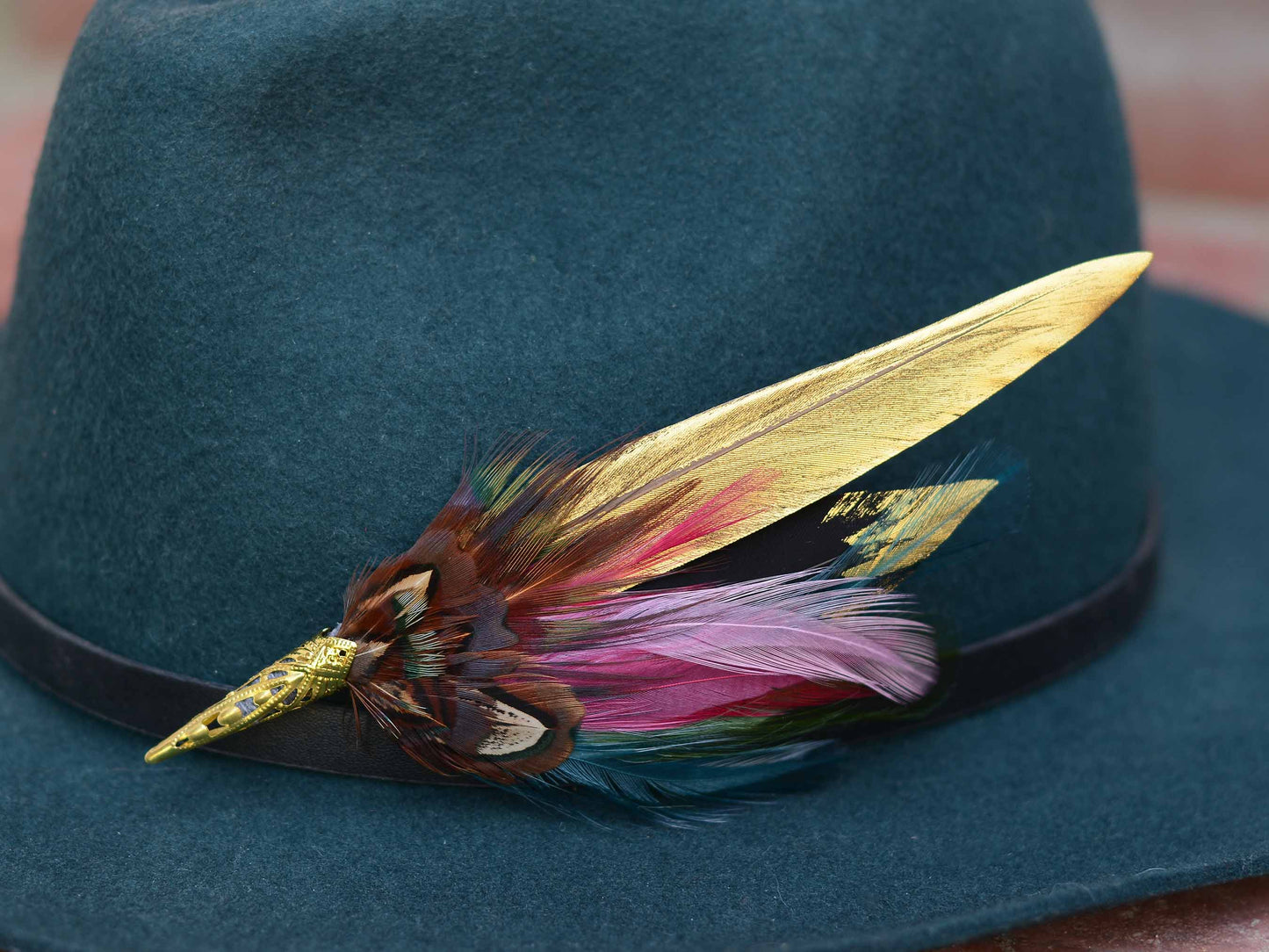 A close-up of a dark teal felt hat with a black band, decorated with a long gold feather hat pin and a cluster of multicolored feathers in pink, purple, brown, and green.