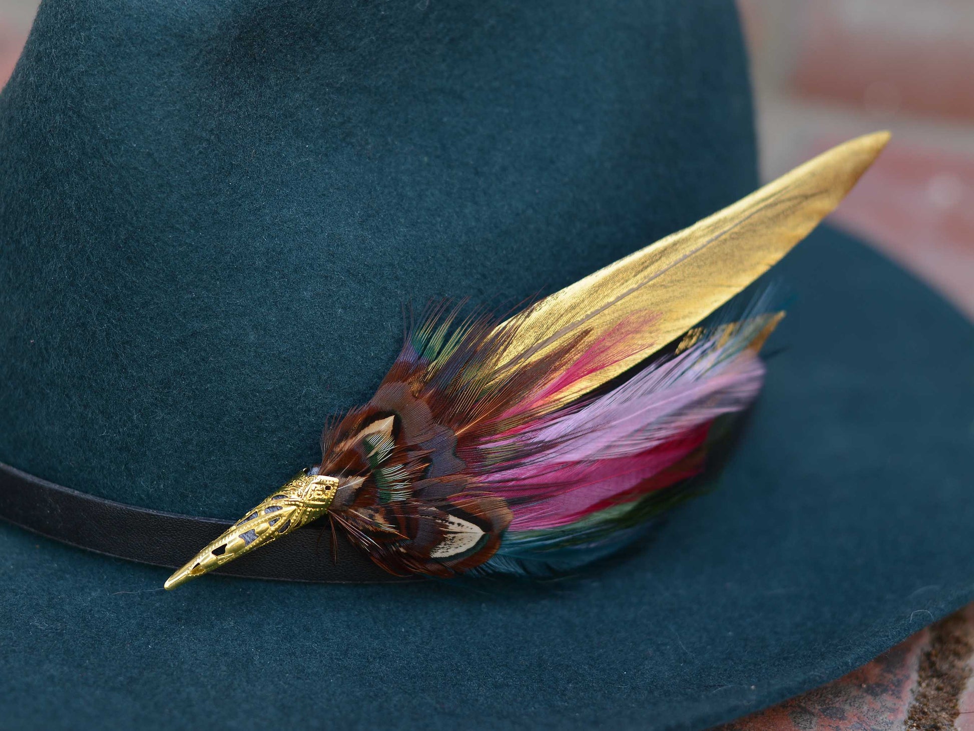 A close-up of a dark teal felt hat with a black band, decorated with a long gold feather hat pin and a cluster of multicolored feathers in pink, purple, brown, and green.