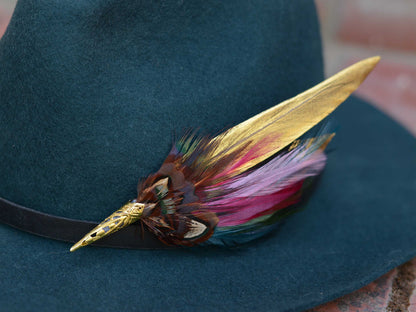 A close-up of a dark teal felt hat with a black band, decorated with a long gold feather hat pin and a cluster of multicolored feathers in pink, purple, brown, and green.