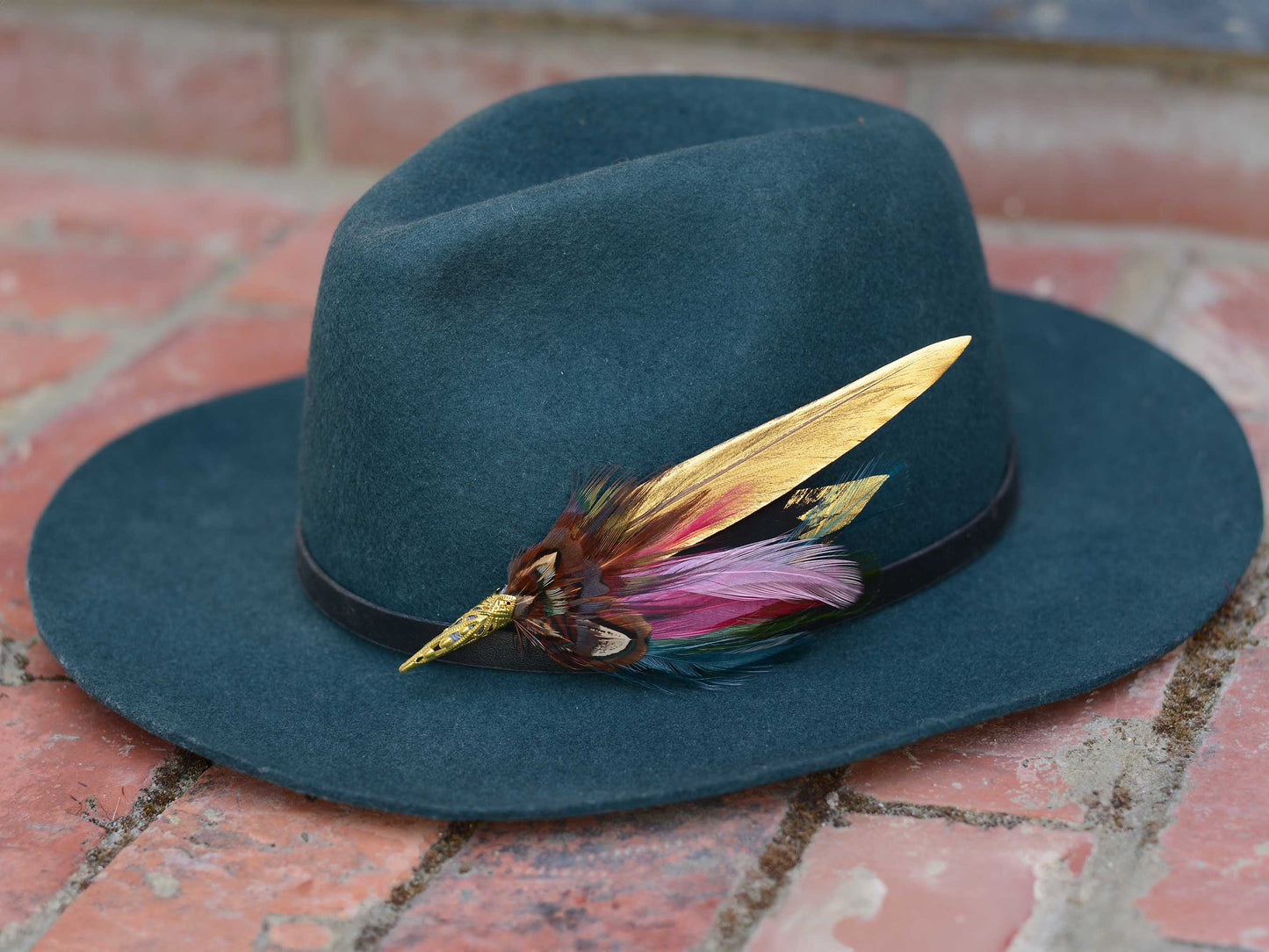 A dark teal felt fedora hat resting on a red brick surface, decorated with a black band and a feather accent featuring gold, pink, and teal feathers.