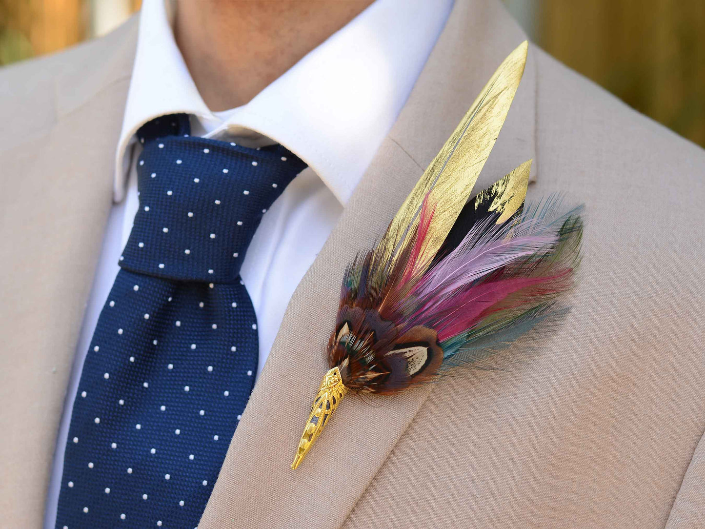 A decorative feather boutonniere pin with gold-toned metal details , handpainted gold feathers, pheasant and multicolored feathers, pinned to a beige suit jacket worn by a man in a white collared shirt and a navy polka-dot tie.