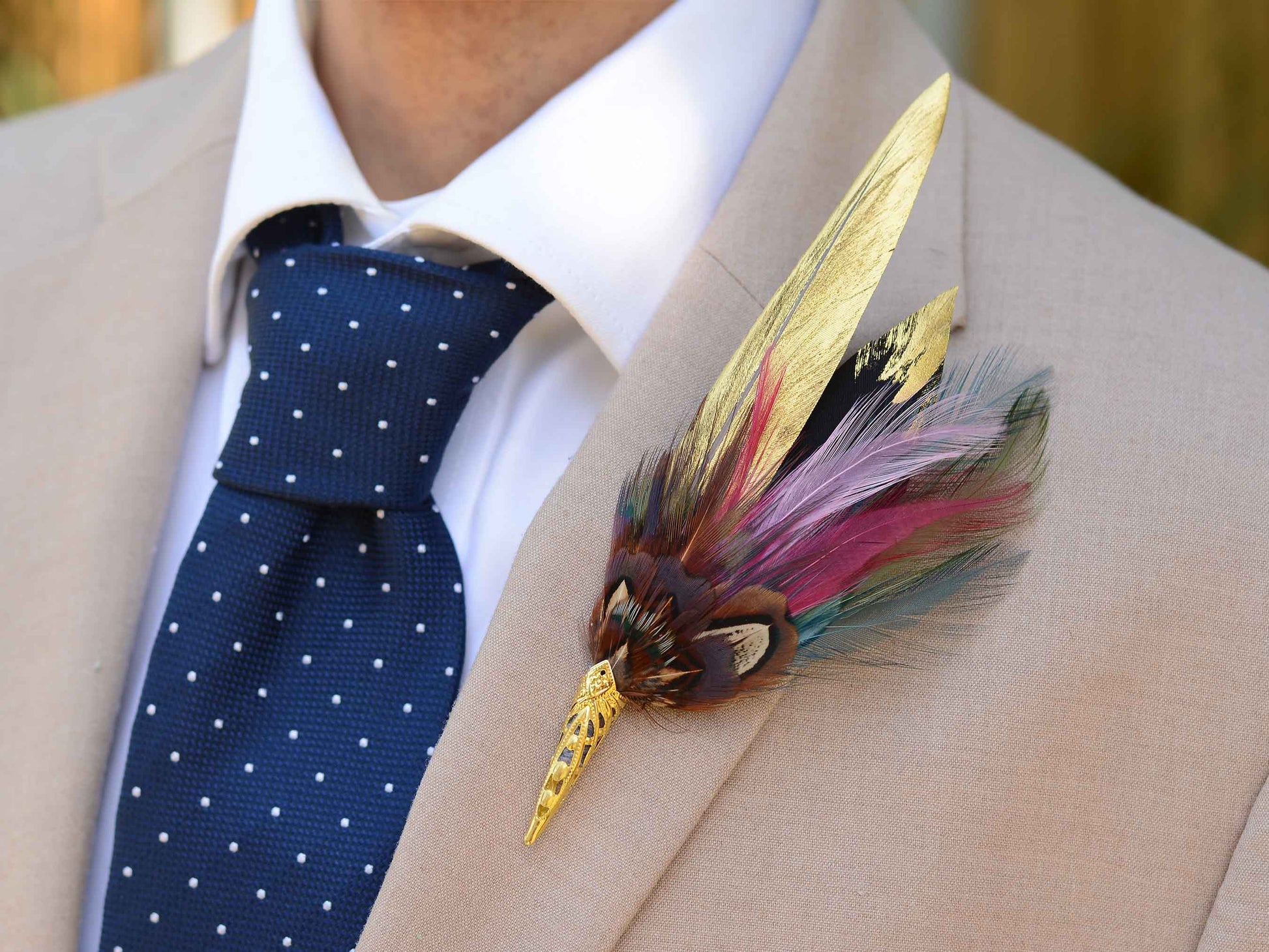 A decorative feather boutonniere pin with gold-toned metal details , handpainted gold feathers, pheasant and multicolored feathers, pinned to a beige suit jacket worn by a man in a white collared shirt and a navy polka-dot tie.