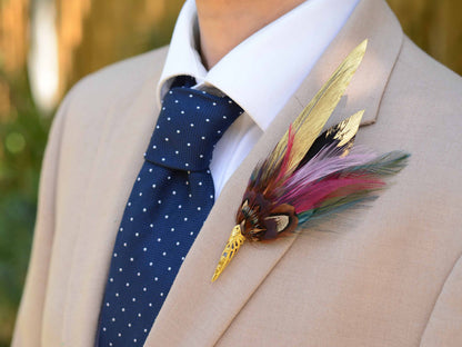 A decorative feather boutonniere pin with gold-toned metal details , handpainted gold feathers, pheasant and multicolored feathers, pinned to a beige suit jacket worn by a man in a white collared shirt and a navy polka-dot tie.