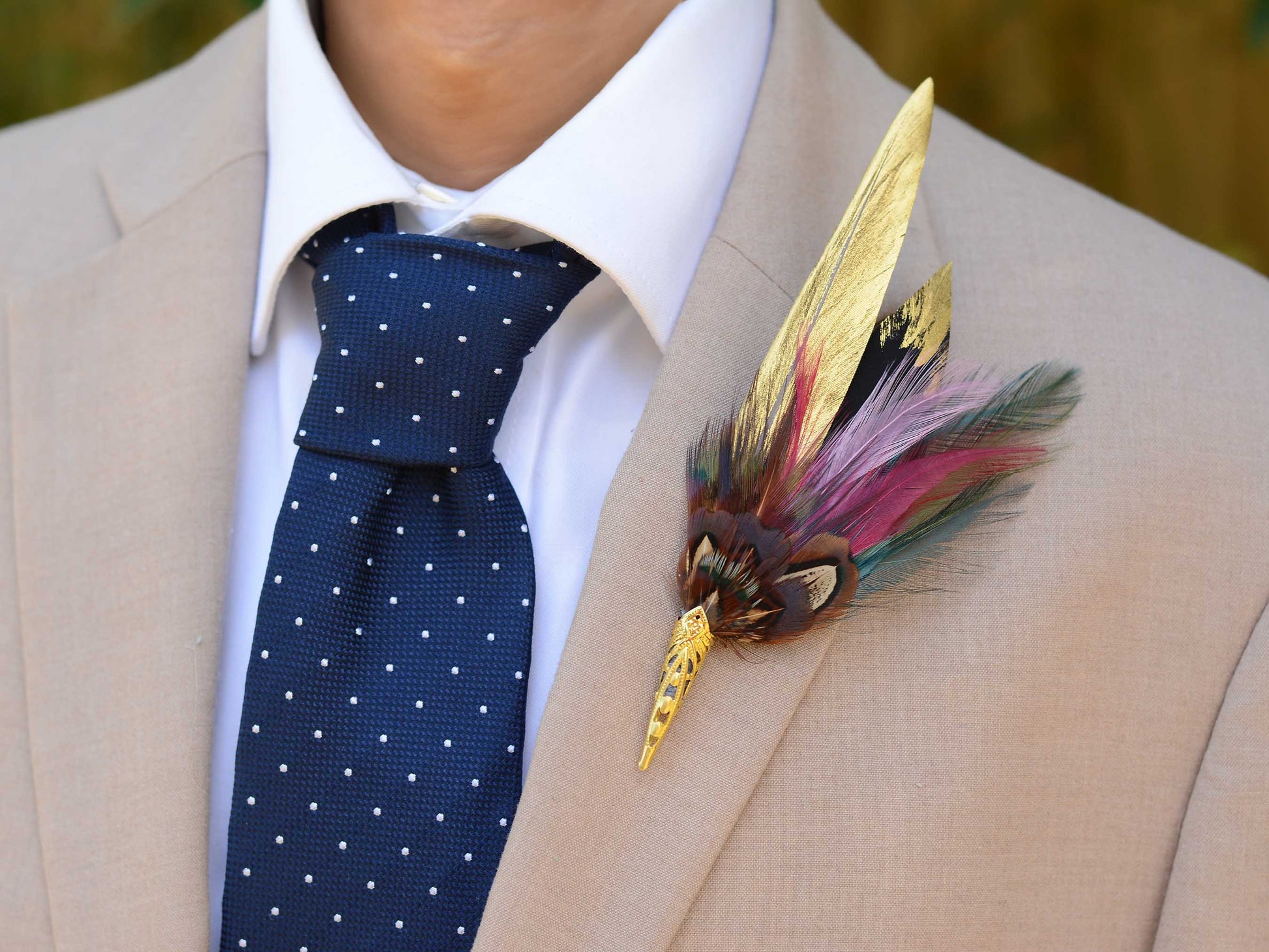 A decorative feather boutonniere pin with gold-toned metal details , handpainted gold feathers, pheasant and multicolored feathers, pinned to a beige suit jacket worn by a man in a white collared shirt and a navy polka-dot tie.