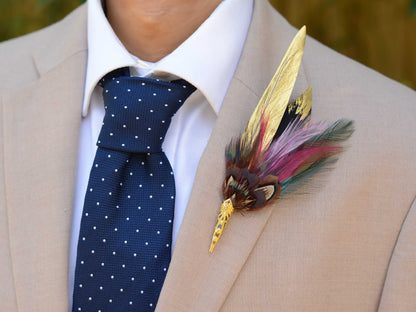 A decorative feather boutonniere pin with gold-toned metal details , handpainted gold feathers, pheasant and multicolored feathers, pinned to a beige suit jacket worn by a man in a white collared shirt and a navy polka-dot tie.