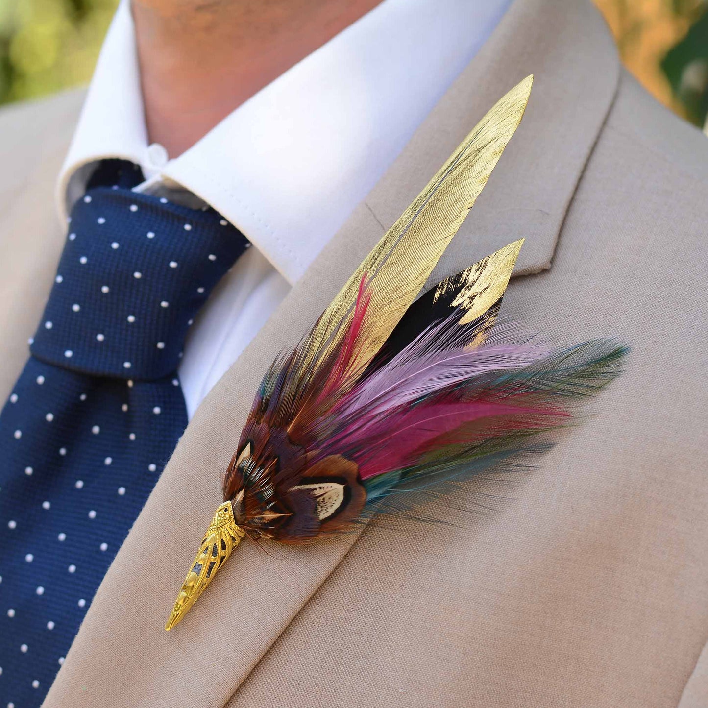 A decorative feather boutonniere pin with gold-toned metal details , handpainted gold feathers, pheasant and multicolored feathers, pinned to a beige suit jacket worn by a man in a white collared shirt and a navy polka-dot tie.