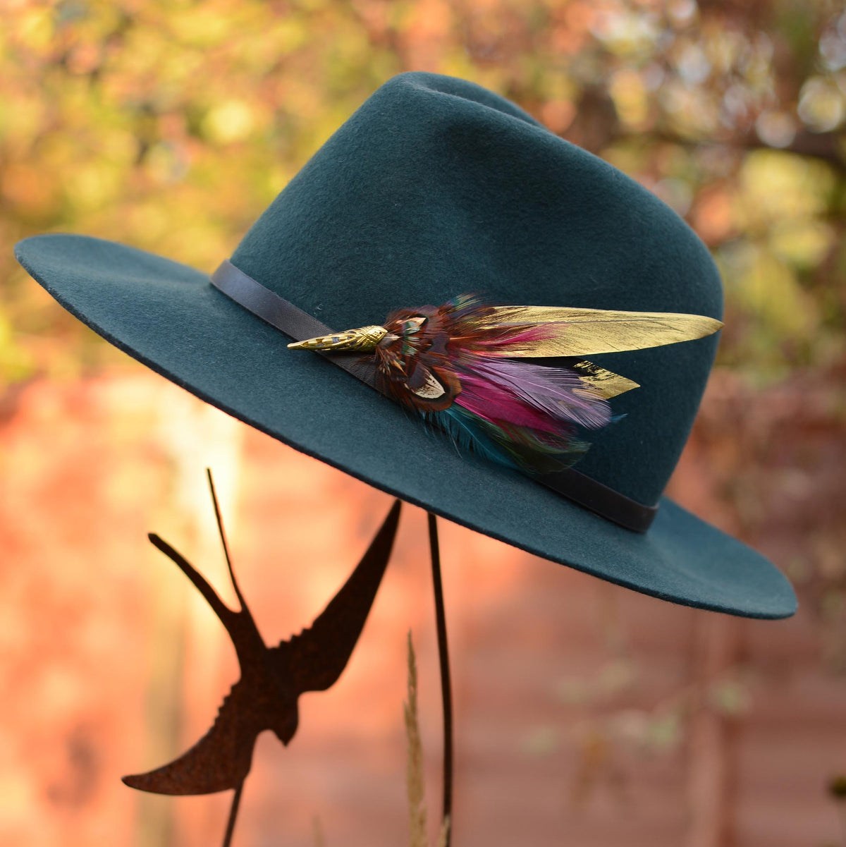 A dark teal felt wide-brim hat with a black band and a decorative feather hat pin brooch in gold, burgundy and teal displayed outdoors against a blurred warm-toned background.