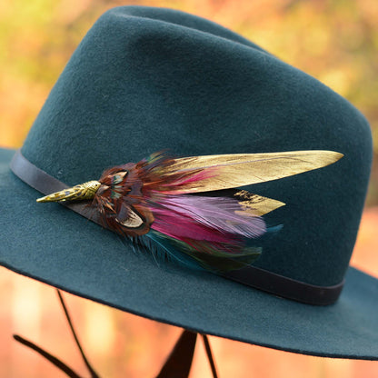 A dark teal felt hat with a black band and a decorative feather accent  pin in gold, brown, magenta, and teal, photographed outdoors against a blurred autumn background.