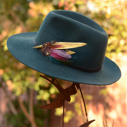 A dark teal felt cowboy hat with a black band and a decorative feather hat pin, displayed on a metal stand against a blurred outdoor background of green foliage and warm sunlight.