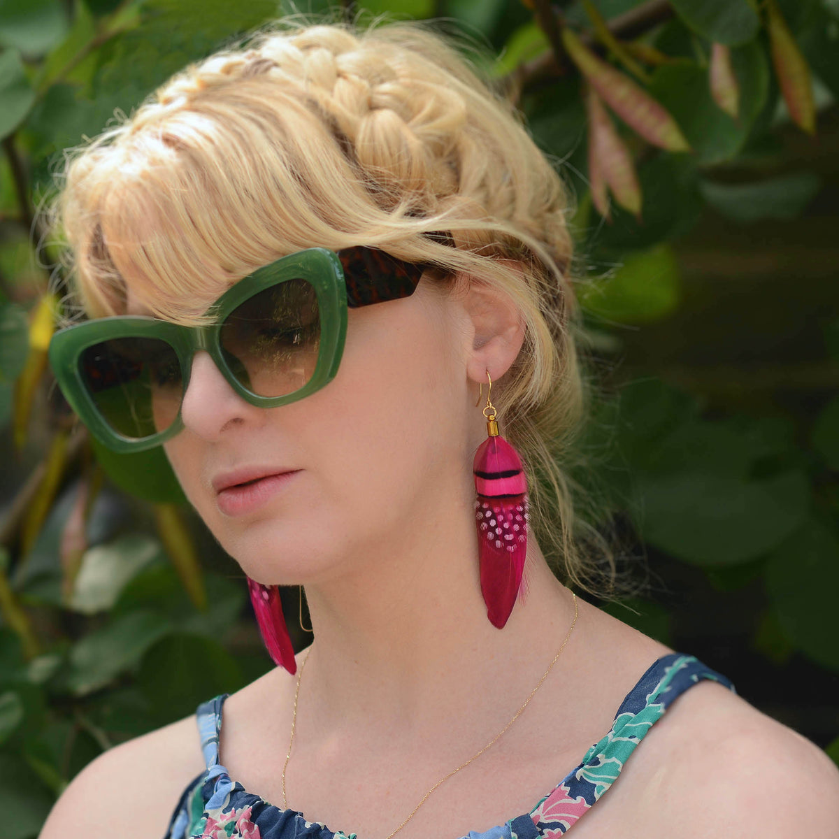 A close-up lifestyle shot of a woman with blonde hair in a braided bun, wearing green cat-eye sunglasses and long magenta boho feather dangle earrings with gold accents, set against a background of green foliage.