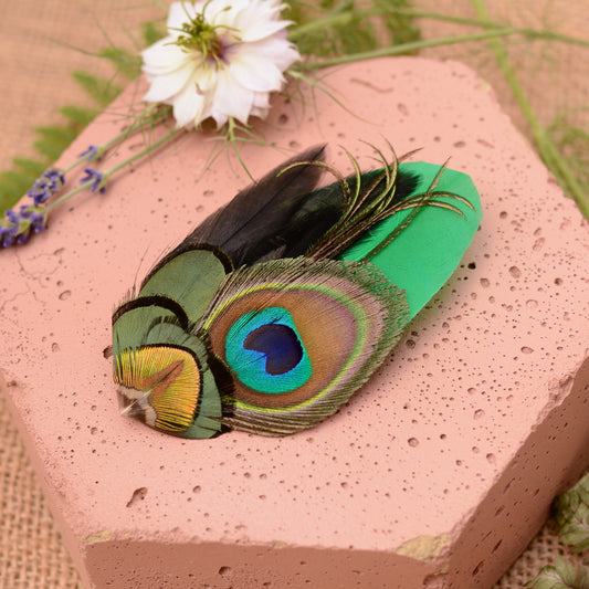 feather hair clip with peacock eye in emerald green gold and blue on pink stone background