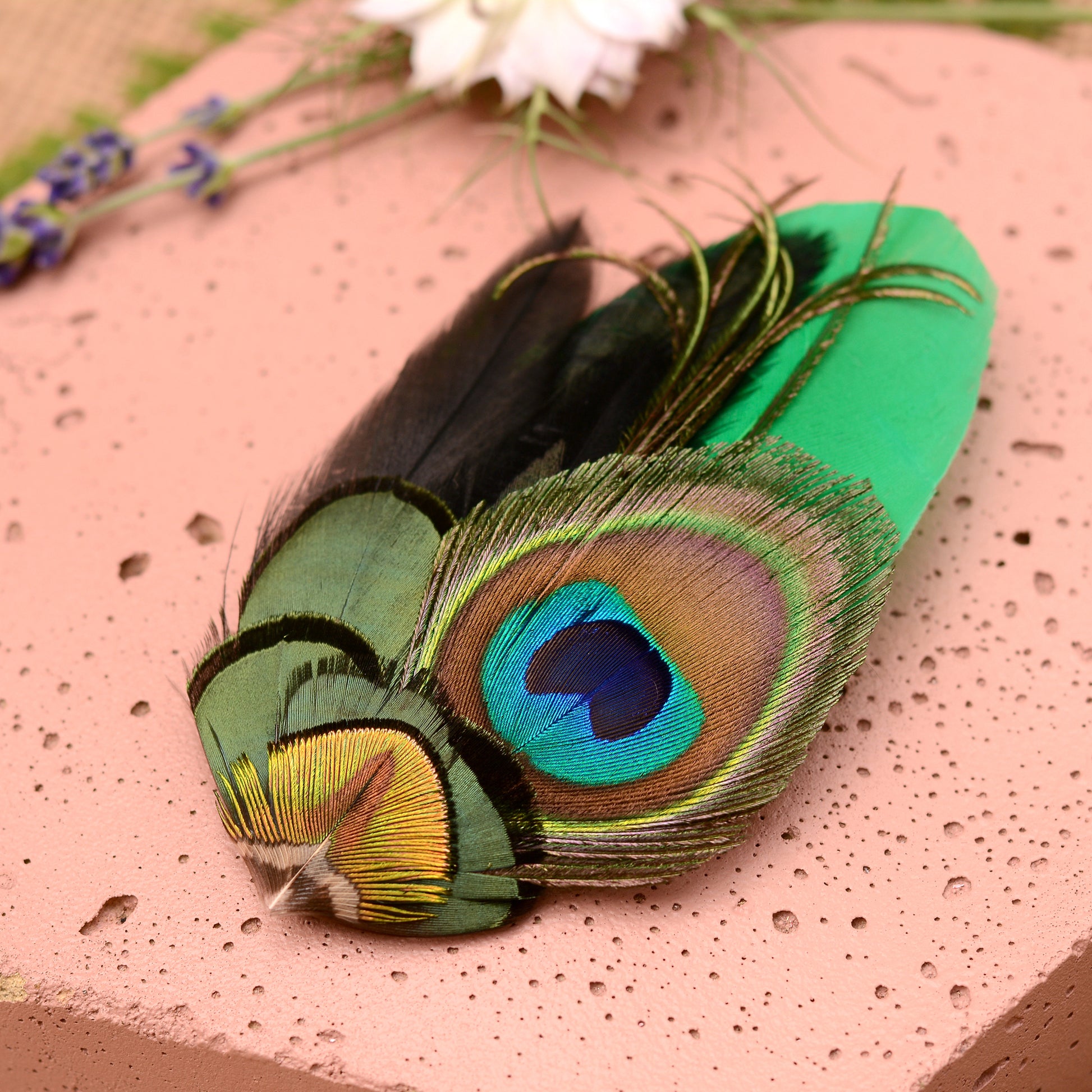 close up of feather hair clip with peacock eye in emerald green gold and blue on pink stone background