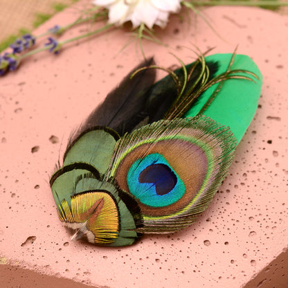 close up of feather hair clip with peacock eye in emerald green gold and blue on pink stone background