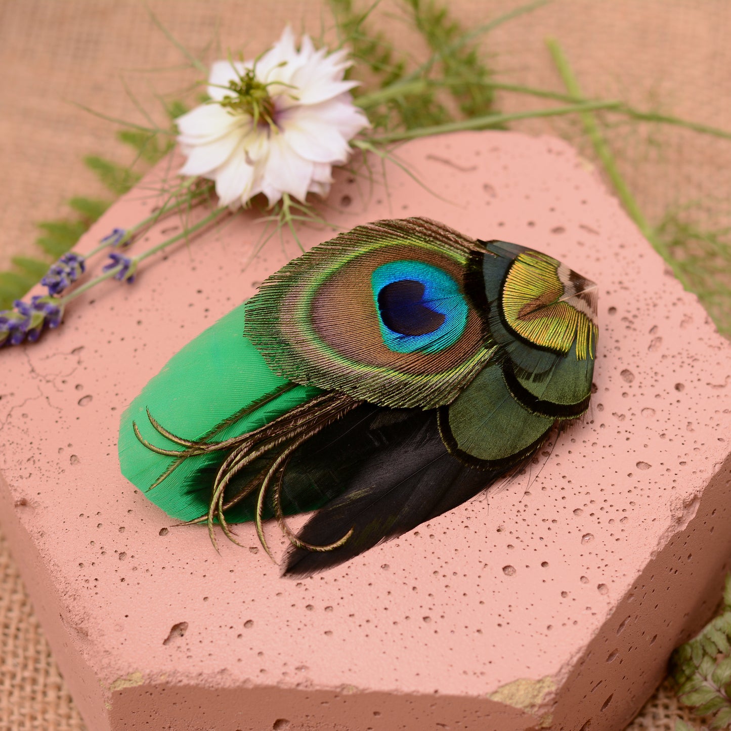 feather hair clip with peacock eye in emerald green gold and blue on pink stone background from side angle