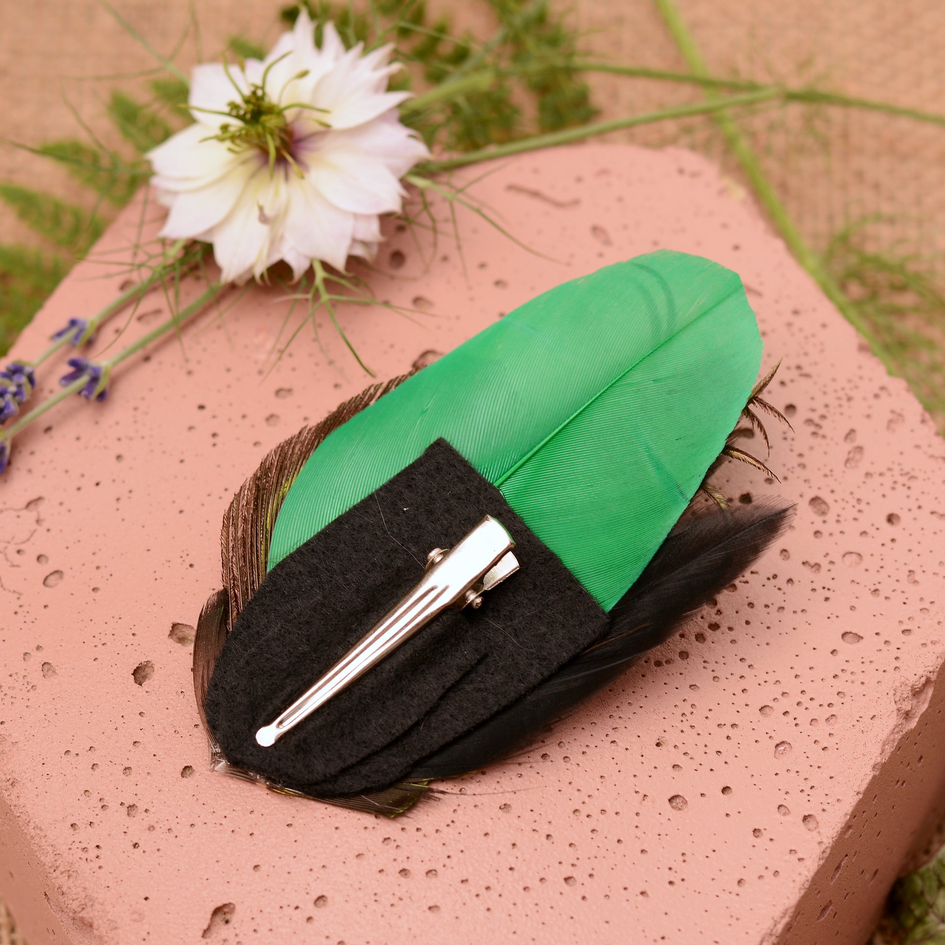feather hair clip with peacock eye in emerald green gold and blue on pink stone background showing back of item and crocodile style clip