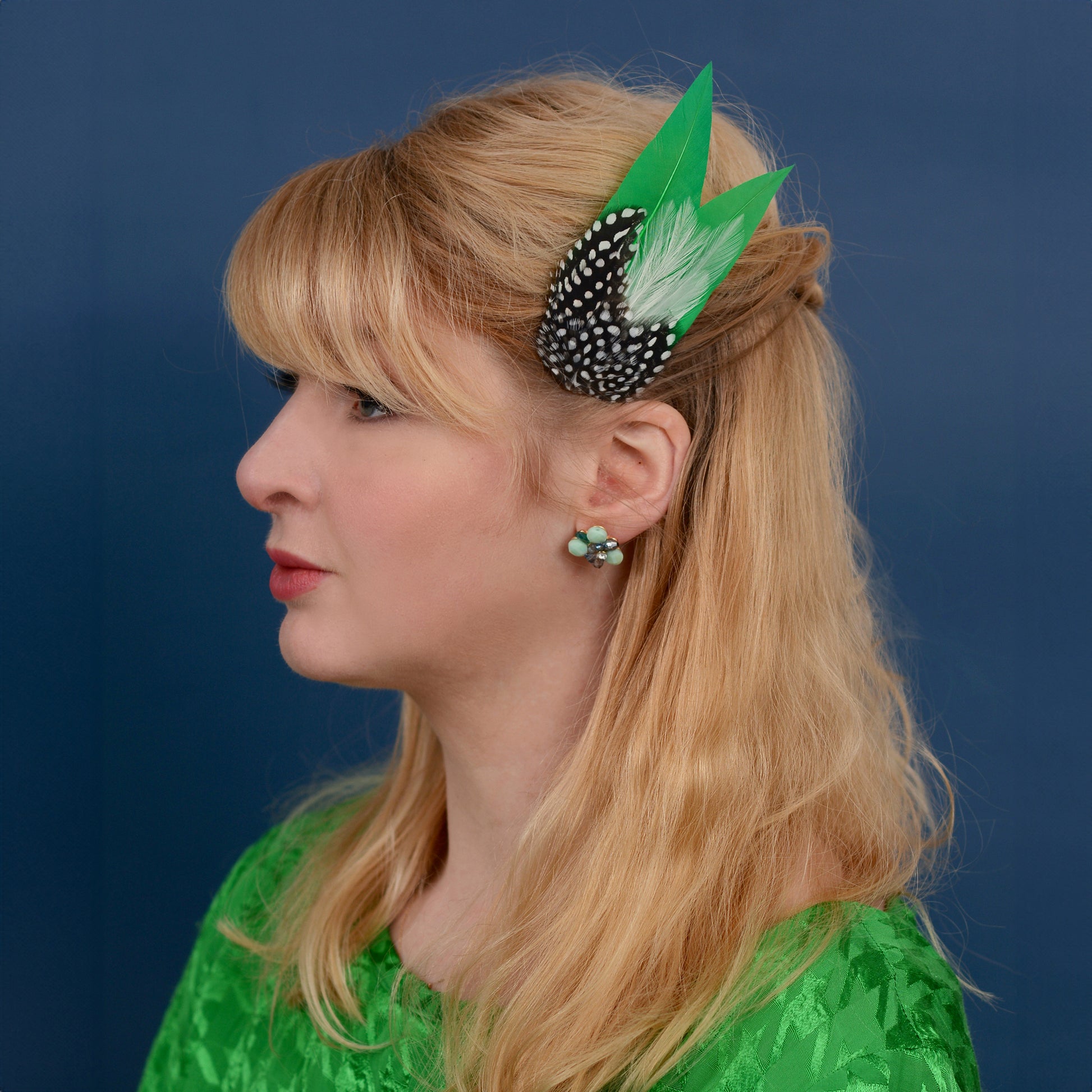 Woman with a green feather hair clip  with polka-dot spotted feathers against a blue background