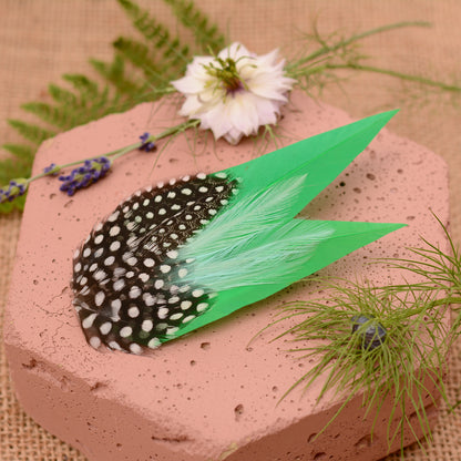 Green feathered hair clip with spotted polka dot feathers on a textured surface with flowers and greenery.