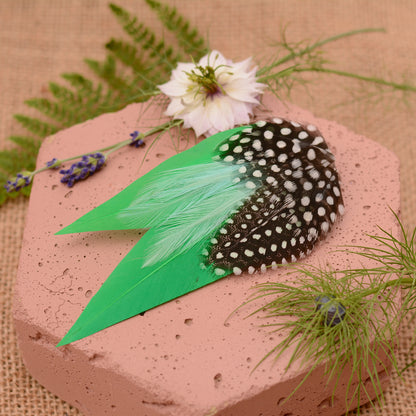 Green feathered hair clip with spotted polka dot feathers on a textured surface with flowers and greenery.