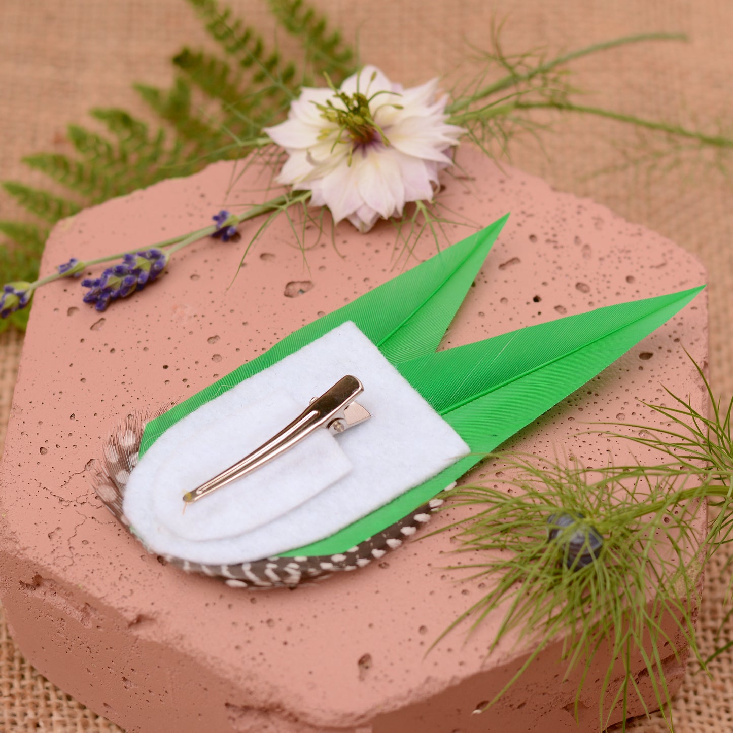 Green feathered hair clip with spotted polka dot feathers on a textured surface with flowers and greenery.