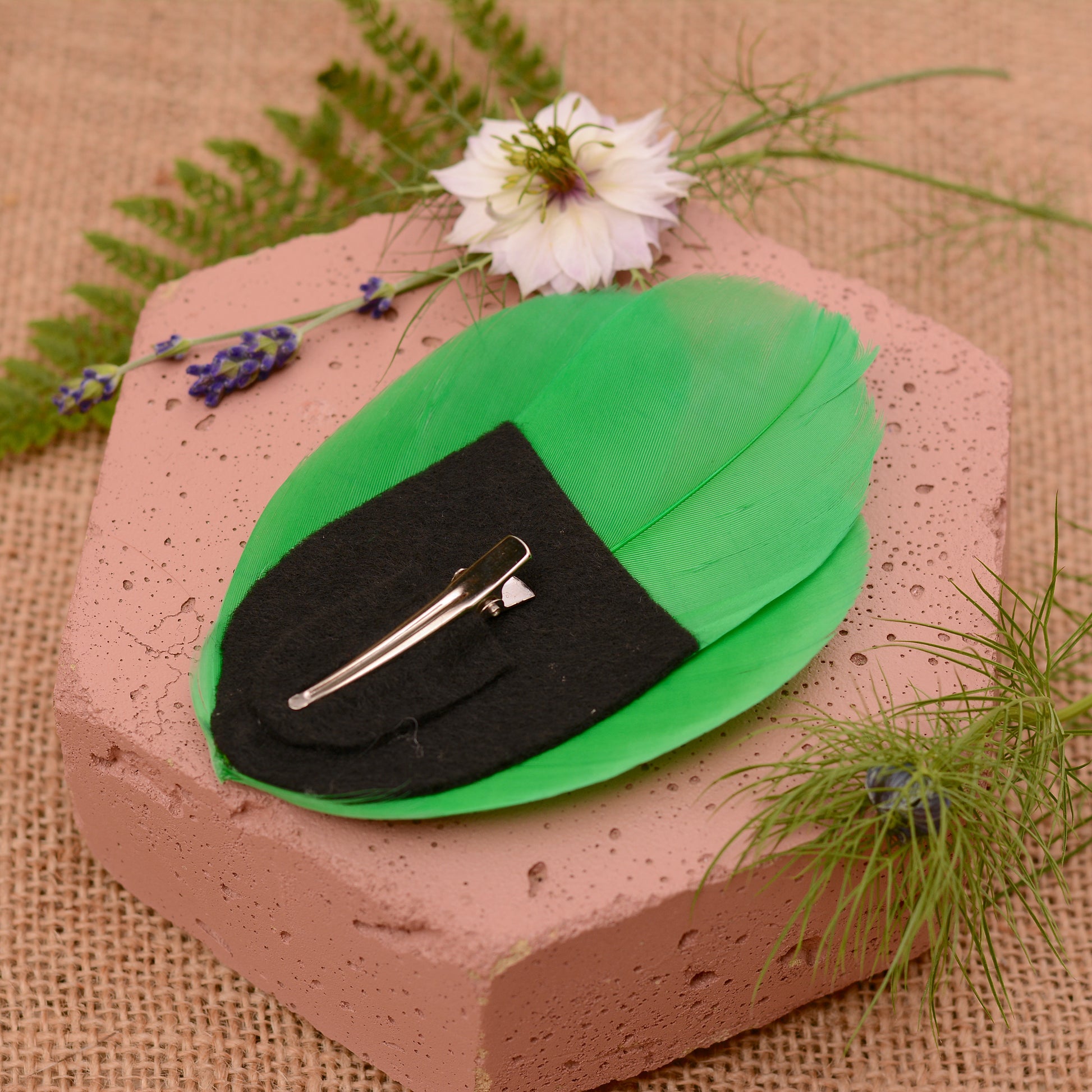 Green and black feathered hair clip on a textured surface with flowers and greenery.