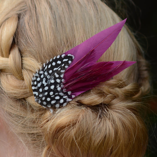 A close-up of a blonde braided hairstyle featuring a decorative hair accessory with black and white spotted feathers and two purple feathers.