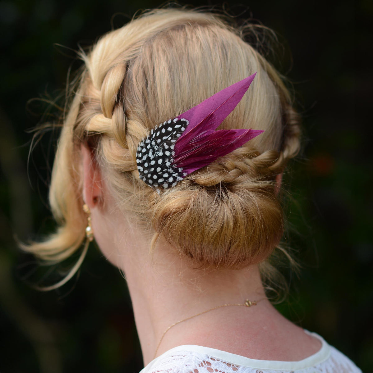 A blonde woman shown from the back wearing a braided bun with a decorative hair accessory featuring purple feathers and  black and white polka dot guinea fowl feathers. Ideal for weddings and formal events.