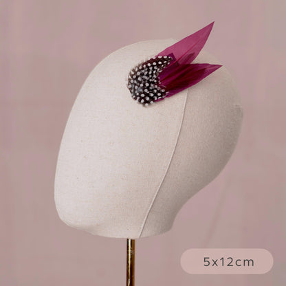 A small decorative hair accessory displayed on a cream fabric mannequin head against a soft pink background. The accessory has two purple feathers and black and white polka dot feathers.