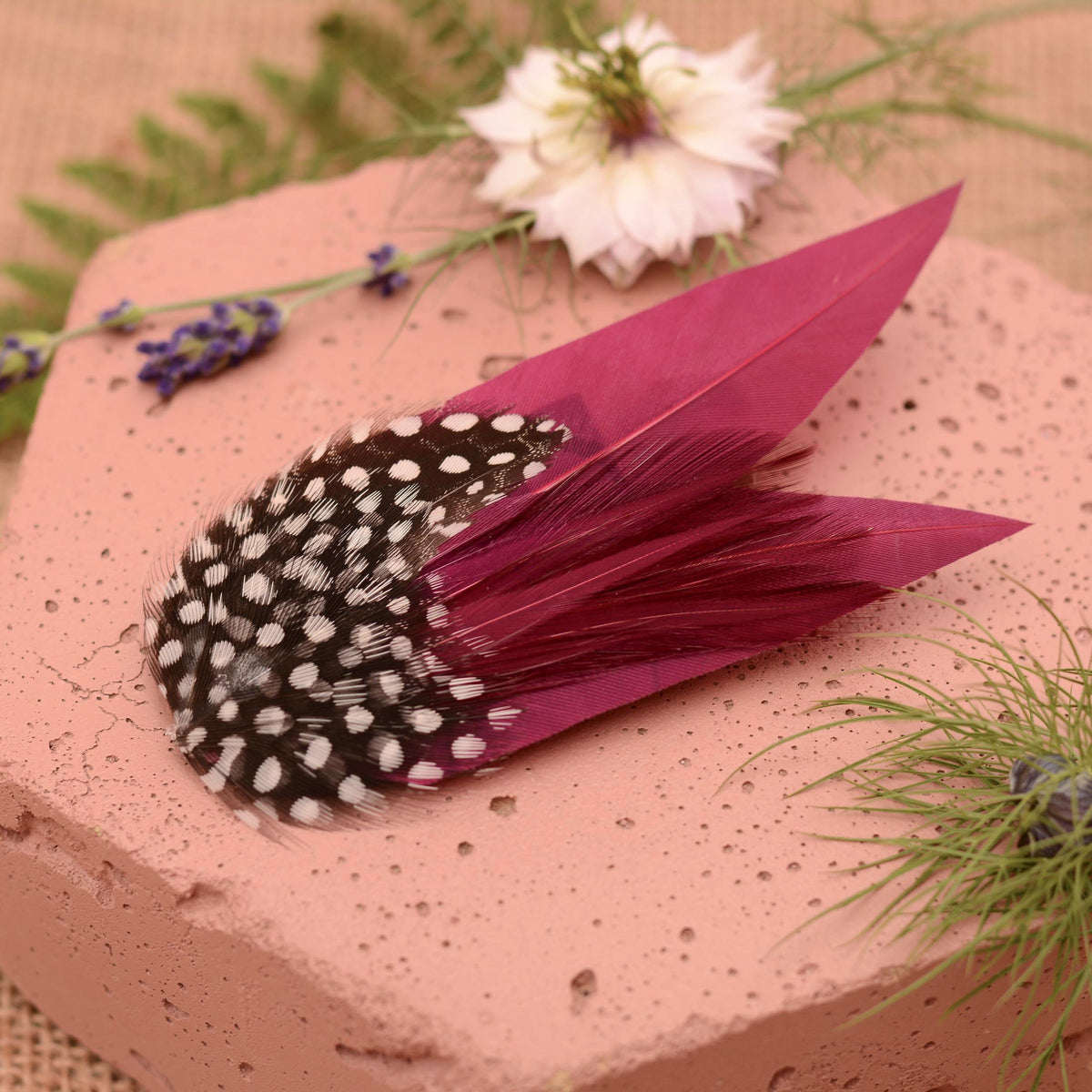 A decorative feather fascinator hair clip in purple with black-and-white spotted guinea fowl feathers, arranged on a pink stone slab with flowers and greenery as styling props.