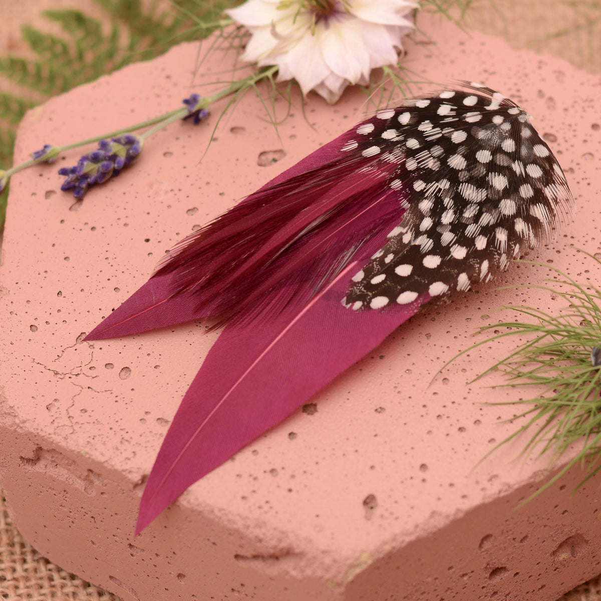 A decorative feather accessory with two long purple feathers and a black-and-white spotted feather section, styled on a terracotta block with flowers and greenery.