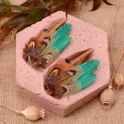 Two decorative wedding feather hair pieces with green, brown, and copper tones arranged on a pink hexagonal block over a burlap surface with dried botanical props.