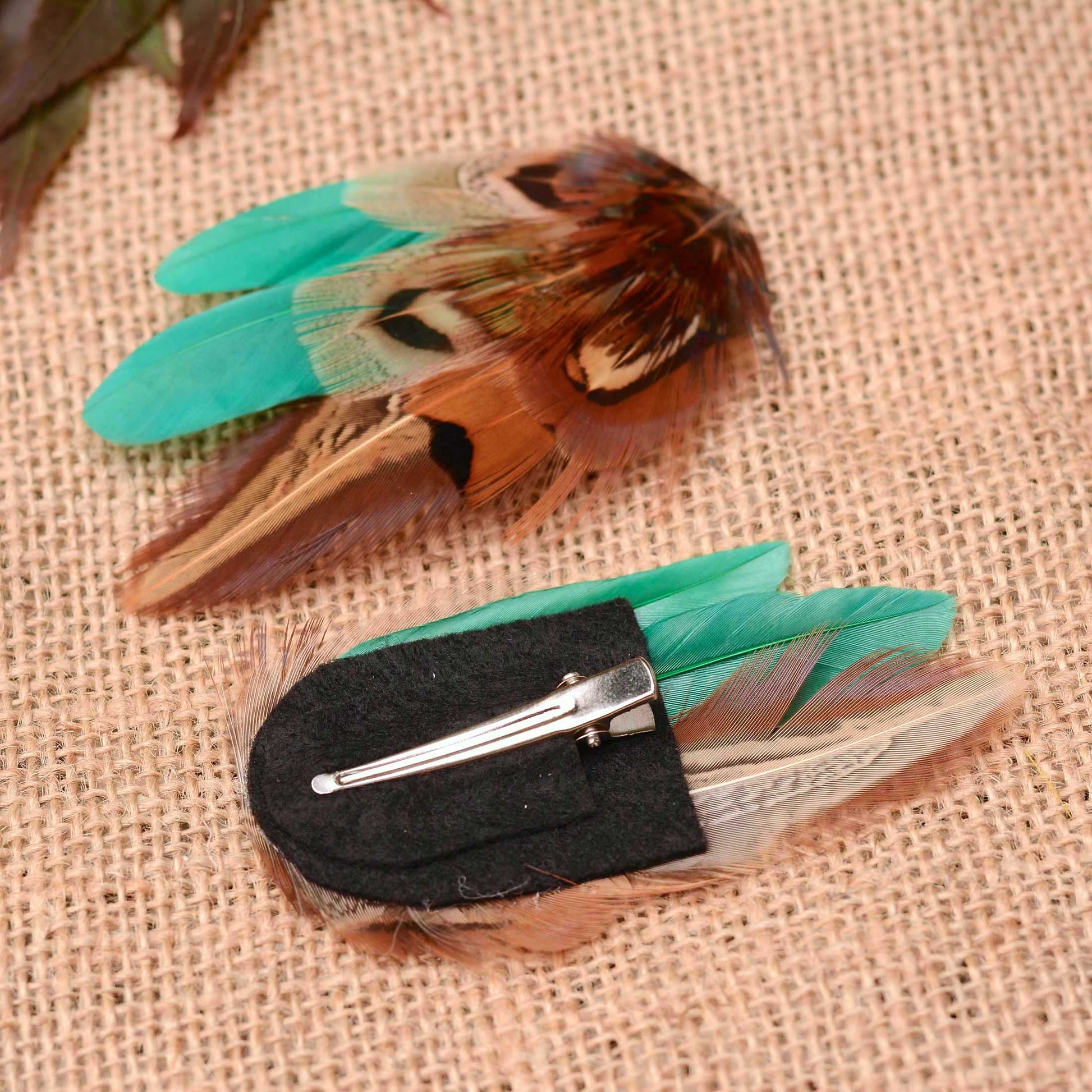 Two feather hair clips on a beige burlap background. One clip features brown, copper and black feathers with green accents, and the other is displayed upside down to show a black felt backing with a silver metal alligator clip  feathers.
