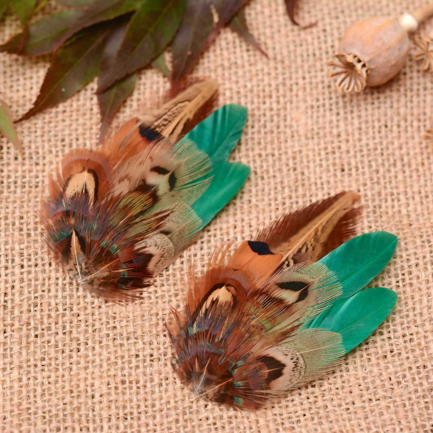 Two decorative pheasant feather clips or hair accessories with brown, copper black, and bright green feathers, arranged on a beige burlap surface with dried leaves and a dried pod in the background.