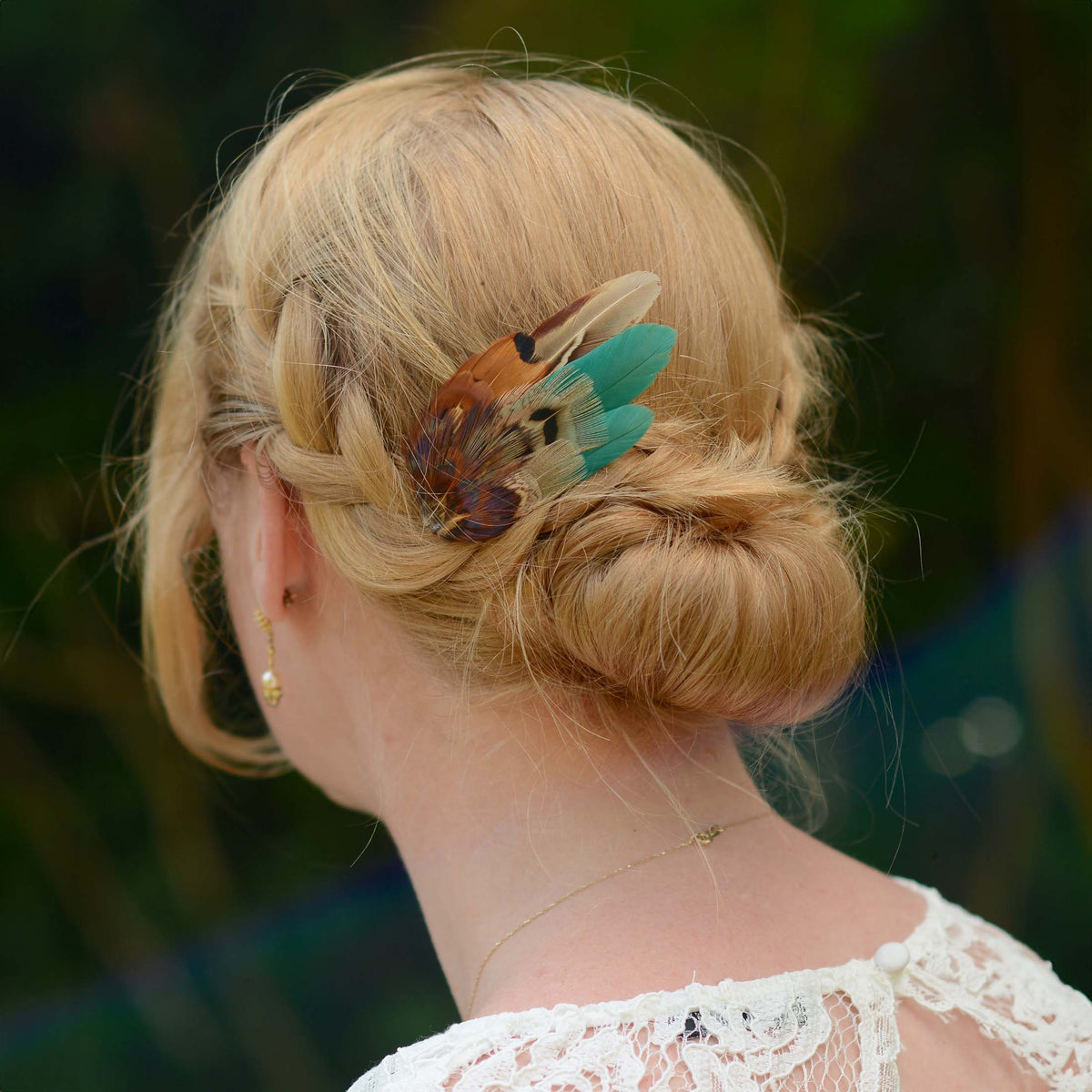 A blonde woman shown from the back wearing a decorative pheasant feather and green hair clip in her low bun, with a blurred outdoor green background.