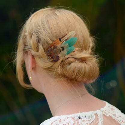 A blonde woman shown from the back wearing a decorative pheasant feather and green hair clip in her low bun, with a blurred outdoor green background.