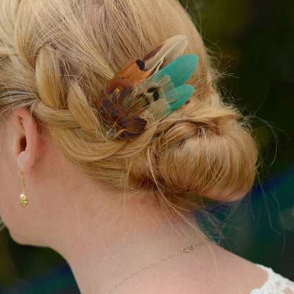 A close-up side view of a woman with blonde hair in a braided bun, wearing a decorative feather hair clip in green copper and brown tones.