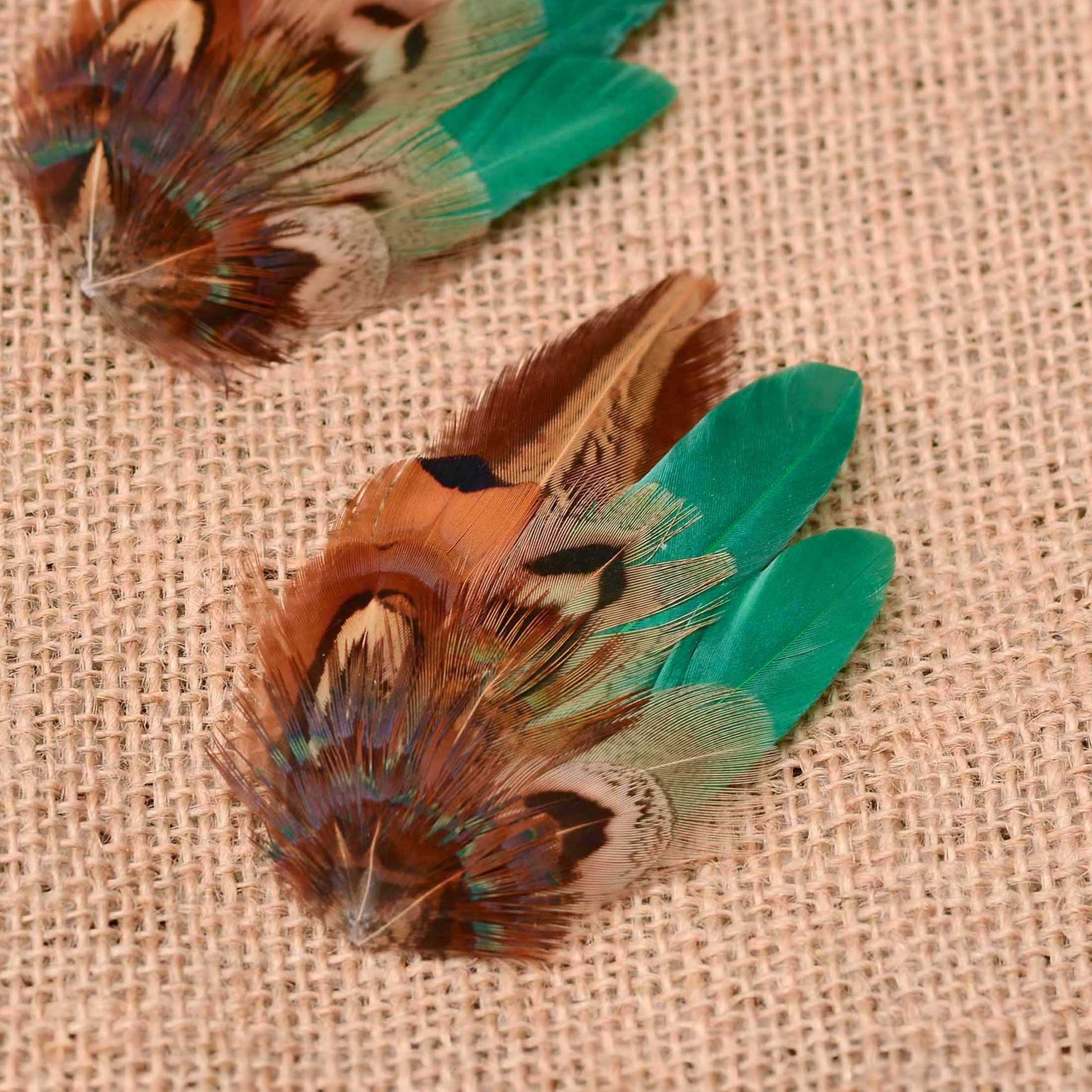 A close-up of a feathered wedding hair clip with brown, copper, and green feathers on a beige burlap background, with a second similar feather visible in the upper left.