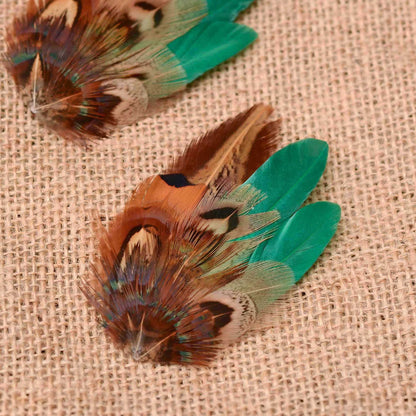 A close-up of a feathered wedding hair clip with brown, copper, and green feathers on a beige burlap background, with a second similar feather visible in the upper left.