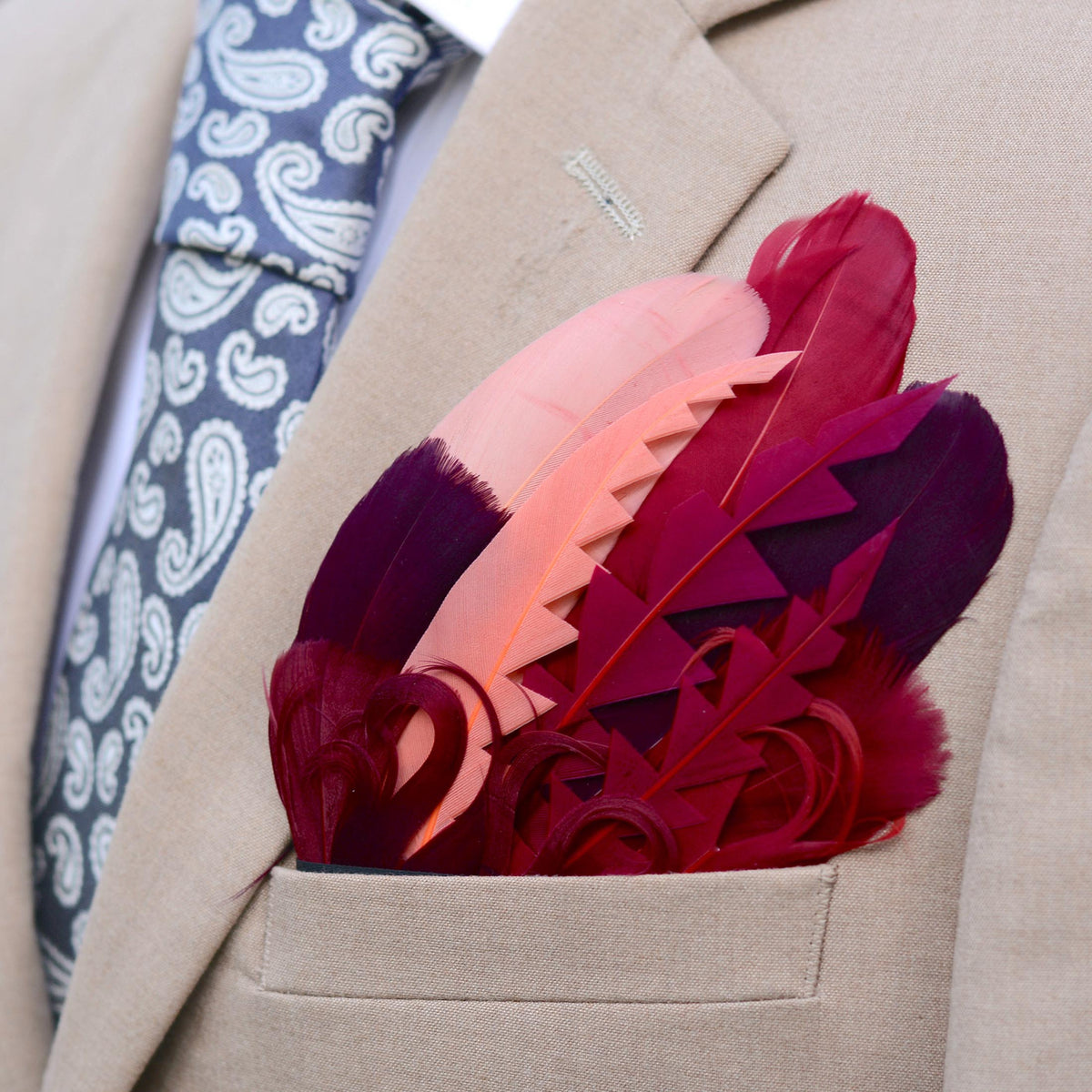A close-up of a beige suit jacket pocket holding a decorative pocket square made of layered feathers in shades of pink, burgundy, and deep purple. A blue paisley necktie is visible at the left side.