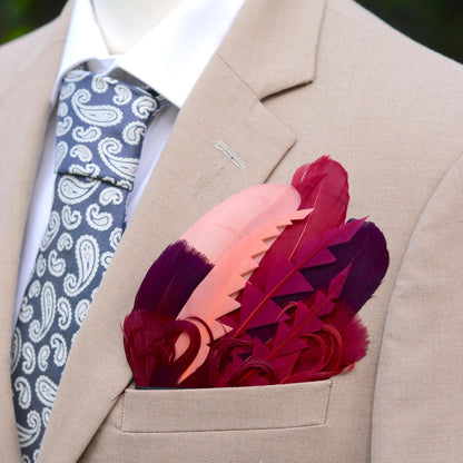A close-up of a beige suit jacket with a white dress shirt and a patterned blue-gray tie, featuring a pocket square made of layered goose  feathers in shades of pink, burgundy, and plum.